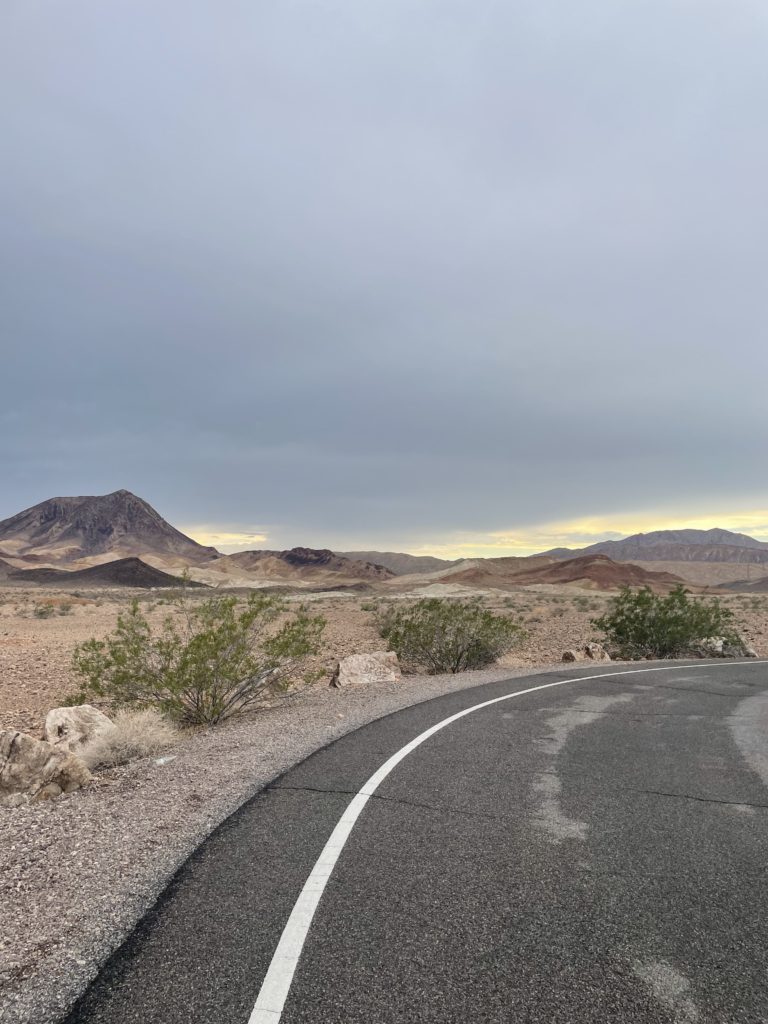 A curved road runs through a barren desert landscape with scattered shrubs and rocky terrain, leading toward distant mountains under a cloudy sky with a hint of sunlight near the horizon.