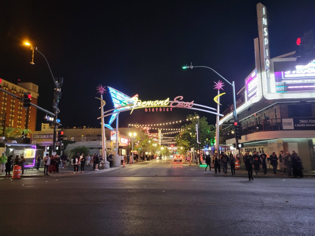 A nighttime street scene at Fremont East District in Las Vegas featuring a neon-lit entrance sign, bright lights, and people walking along the sidewalks under a dark sky.