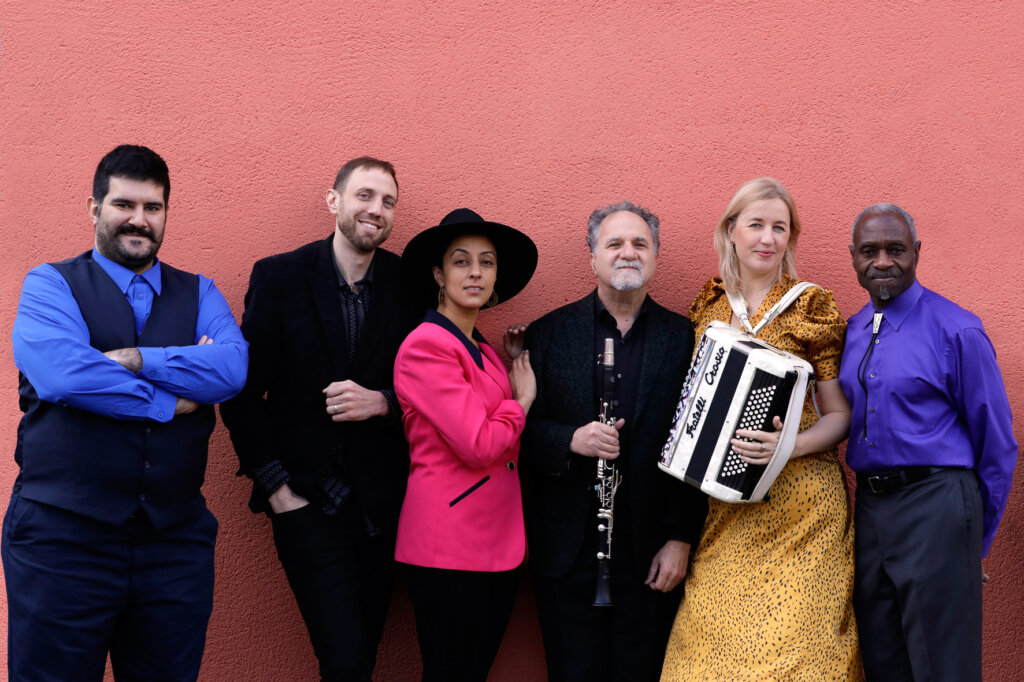Six people stand in a row against a coral wall. One woman in yellow holds an accordion, another in a pink blazer and black hat leans on a man holding a clarinet. The group wear colorful, semi-formal outfits and smile at the camera.