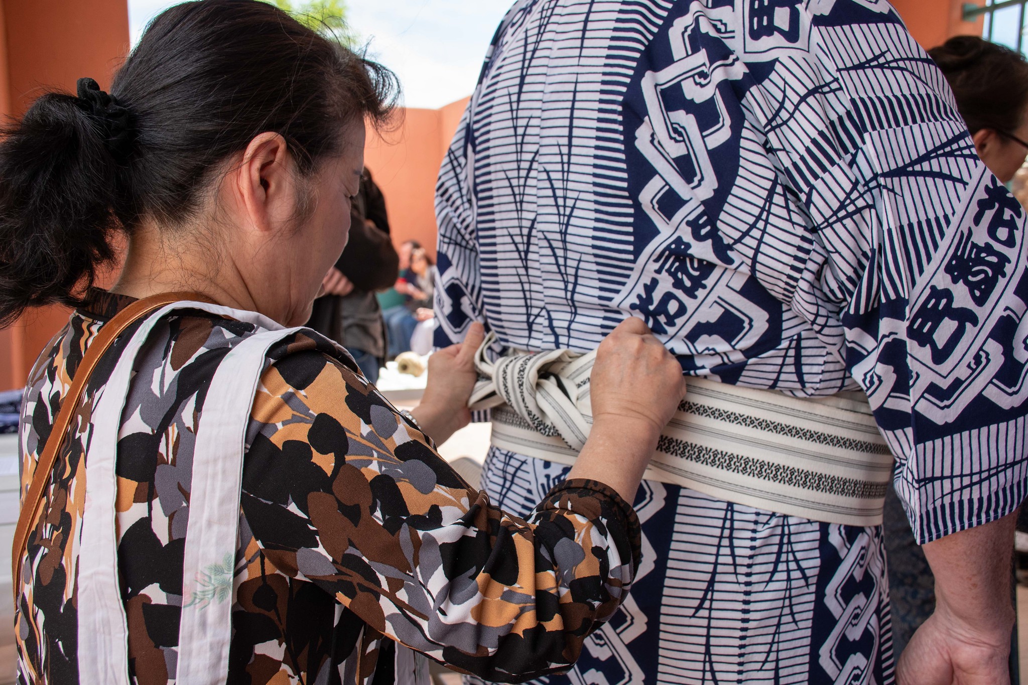A woman ties the obi sash on another person wearing a traditional patterned Japanese yukata, focusing on the hands and fabric details. Other people are blurred in the background.