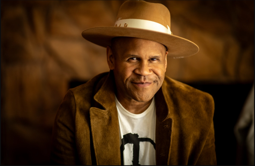 A man wearing a tan wide-brimmed hat and brown suede jacket smiles warmly at the camera, with a blurred brown background.