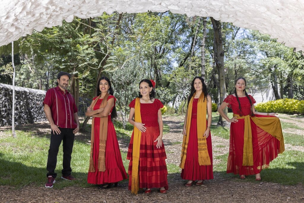 Five people pose outdoors under a white arch. Four women in red dresses with yellow scarves stand together, while one man in a red shirt and black pants stands to the left. Green trees and sunlight fill the background.