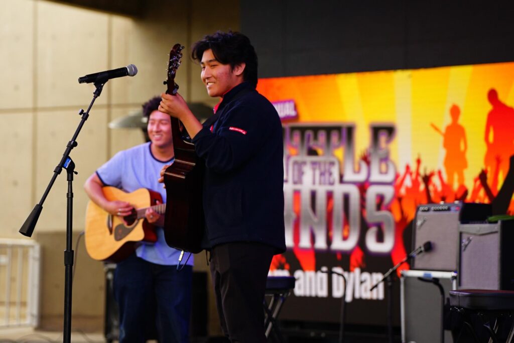Two young men play acoustic guitars and smile on stage during a Battle of the Bands event. Bright lights and a colorful event banner are visible in the background. A microphone stands in front of them.