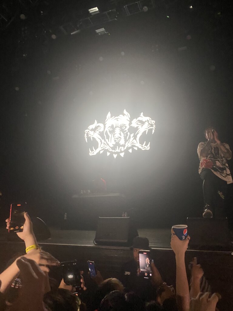 A performer stands on stage with a microphone, illuminated by bright light. Behind them is a large, glowing image of a wolf with three heads. Audience members hold up phones and a Pepsi cup in the foreground.
