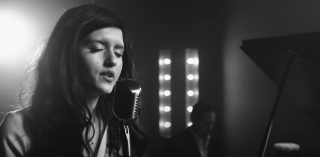 A woman with long dark hair sings into a vintage microphone in a black and white setting. Soft lighting and blurred background reveal a piano and another person in the distance, evoking a classic, moody atmosphere.