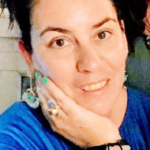 A woman with dark hair pulled back, wearing a blue top, bracelets, rings, and earrings, rests her face on her hand and smiles softly at the camera.