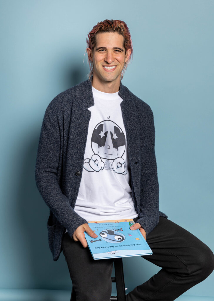 A smiling person with pink hair, wearing a blue cardigan and a white graphic t-shirt, sits on a stool against a light blue background and holds a children’s book titled The Adventures of My Lost Sock.