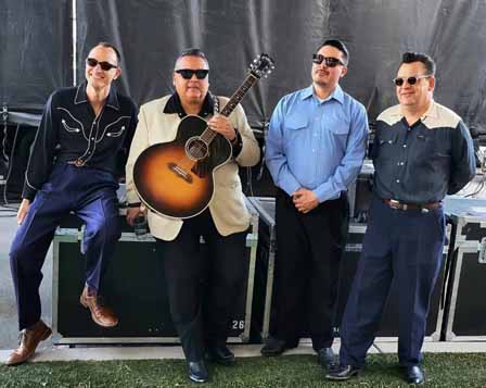 Four men in vintage-style outfits and sunglasses pose together; one holds an acoustic guitar. They are standing outdoors in front of stage equipment.