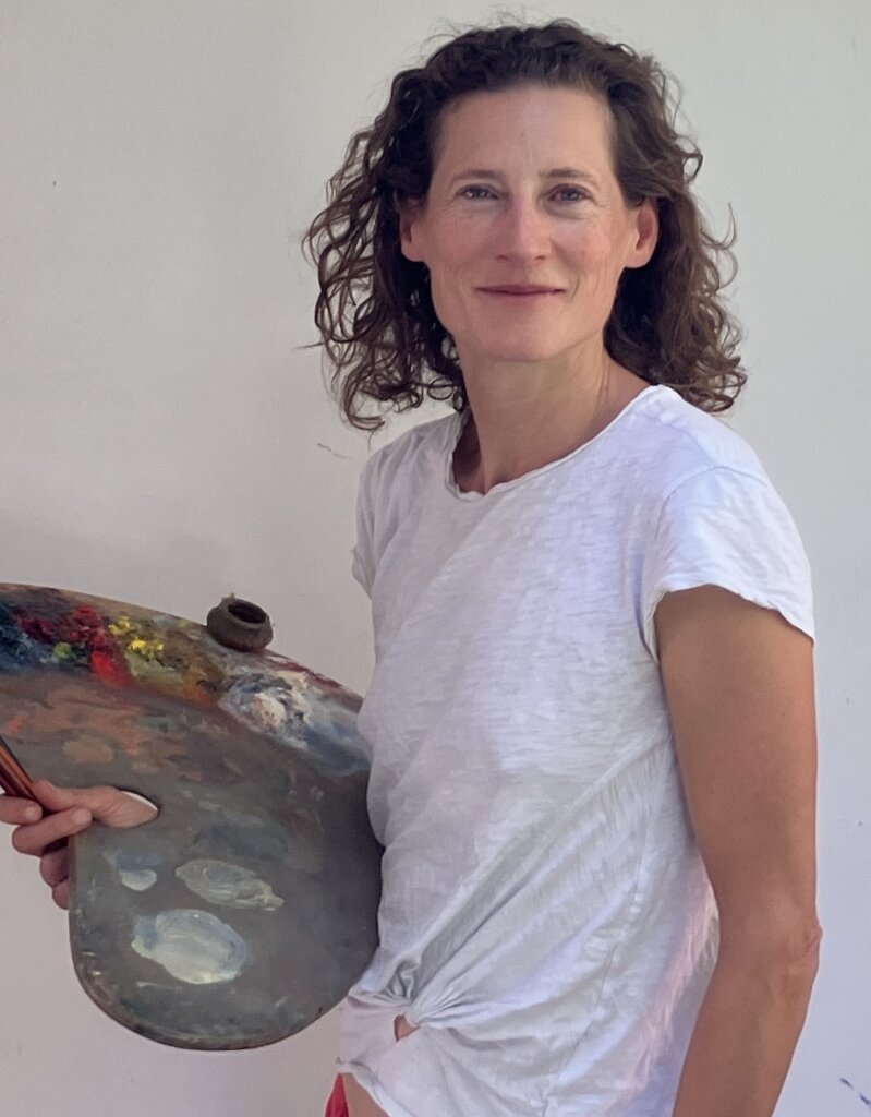 A woman with curly brown hair, wearing a white t-shirt, holds a large artist’s palette with smeared paint. She stands against a plain light-colored background and looks at the camera, smiling softly.