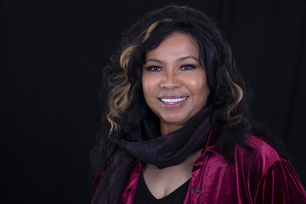 A woman with long dark wavy hair and light highlights smiles at the camera. She is wearing a red velvet top and a black scarf, with a black background behind her.
