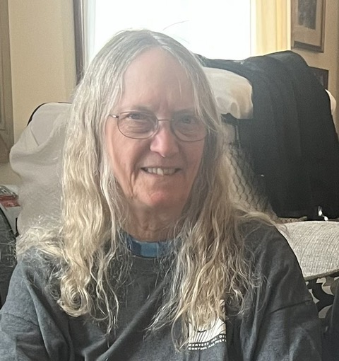 Terrye Kocher: An older woman with long gray hair and glasses is smiling while sitting indoors. She is wearing a dark T-shirt and is in a cozy, softly-lit room with a window and furniture in the background.