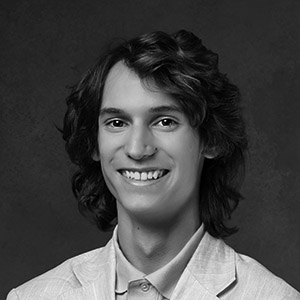 A young person with long wavy hair, wearing a light-colored blazer and collared shirt, smiles at the camera against a dark, plain background. The image is in black and white.