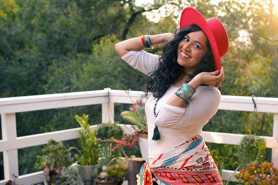 Smiling woman in a red hat poses on a balcony with plants, wearing colorful accessories and a patterned scarf, with trees and sunlight in the background.