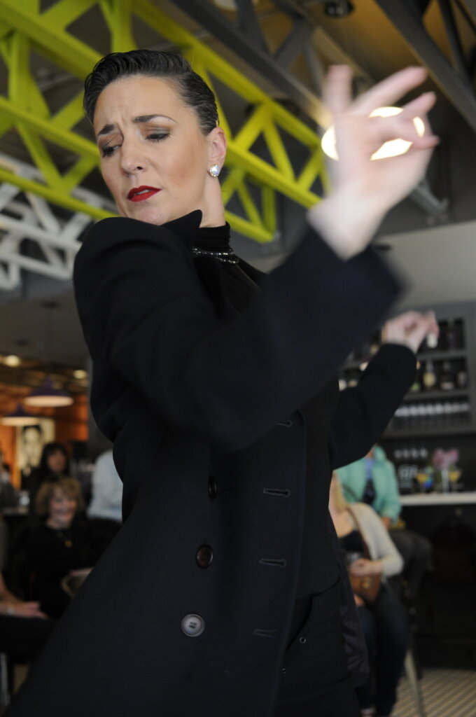A woman wearing a black outfit performs a dramatic dance gesture with her eyes closed and hand raised, in an indoor setting with people seated in the background.