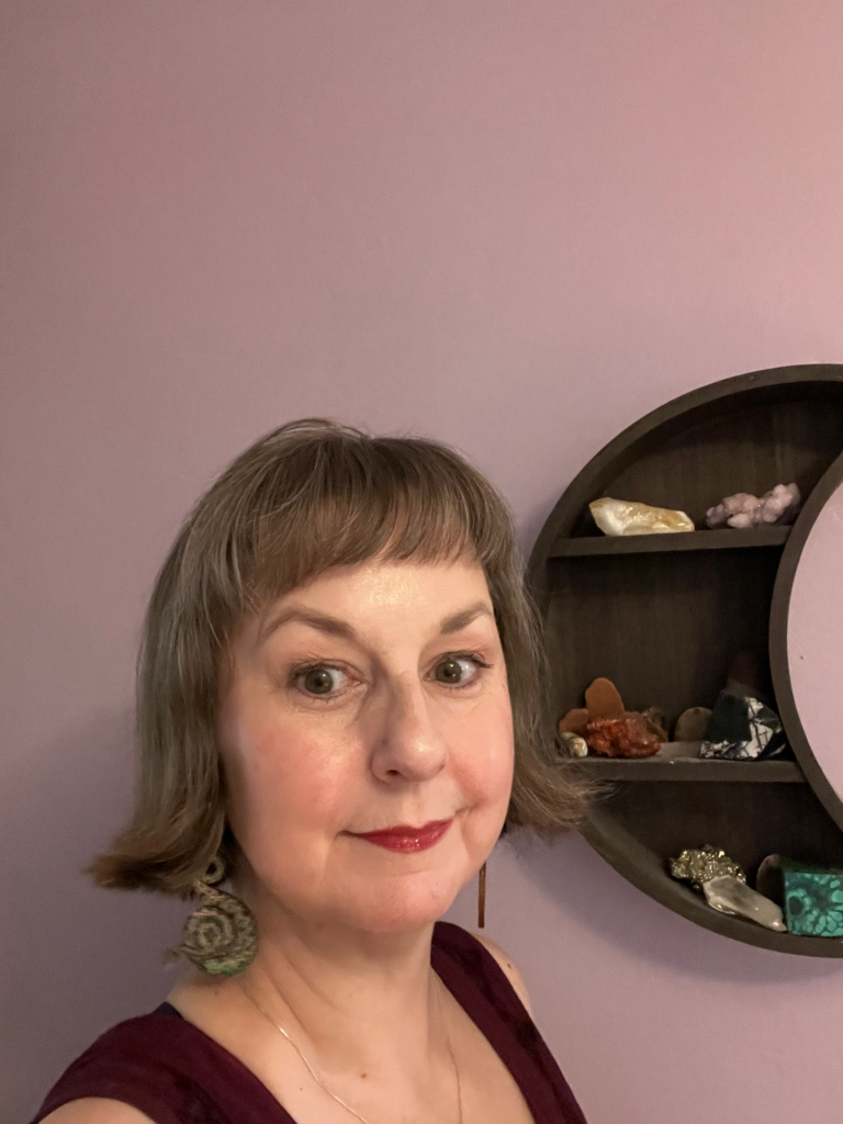 A woman with short brown hair and red lipstick smiles slightly, standing in front of a purple wall. Behind her is a circular shelf displaying various crystals and stones.