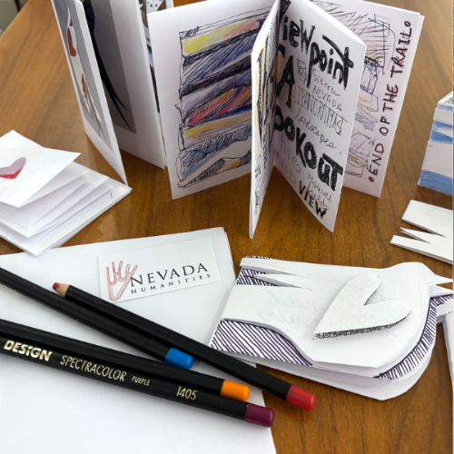 Close-up of handmade mini books with illustrated pages, colored pencils, and a Nevada Humanities card on a wooden table. Some book pages feature drawings, text, and cut-out shapes.