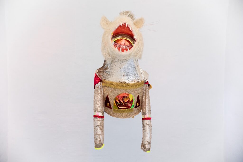 A decorative, handmade puppet of a lion-like creature with a silver body, a wicker-textured torso featuring a cobra emblem, and an open, wide-toothed mouth.