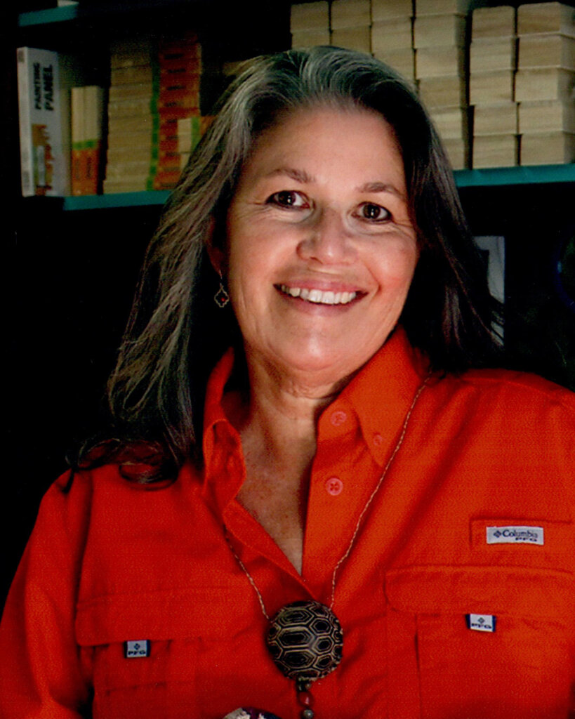 A smiling woman with long gray hair wearing an orange button-up shirt and a large pendant necklace, standing indoors with shelves of books in the background.