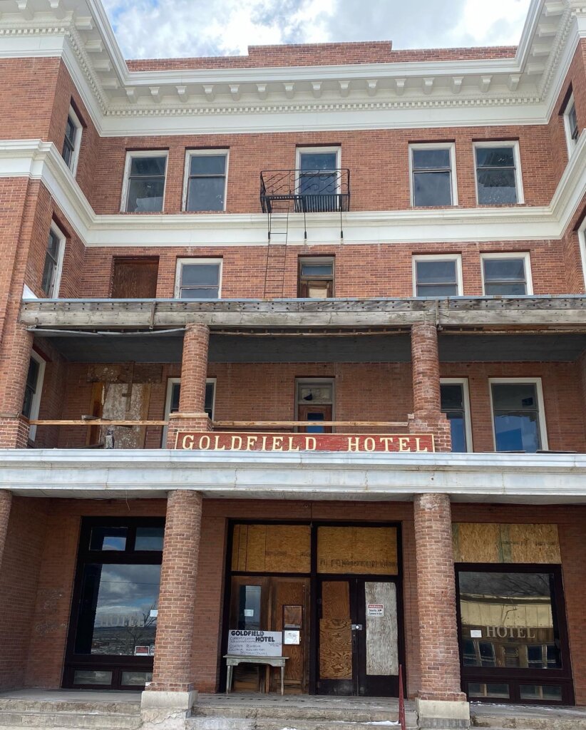 A three-story red brick building with boarded-up windows and doors. A partially damaged sign reads GOLDFIELD HOTEL. The upper balcony and entrance show signs of disrepair.