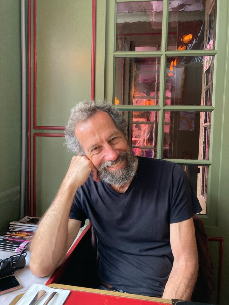 A man with curly gray hair and a beard, wearing a black t-shirt, sits at a table and smiles with his head resting on his hand. Behind him is a window reflecting pink and orange light.