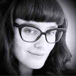 Black and white close-up portrait of a person with wavy hair, blunt bangs, wearing large dark-rimmed glasses and a septum piercing, softly smiling at the camera.