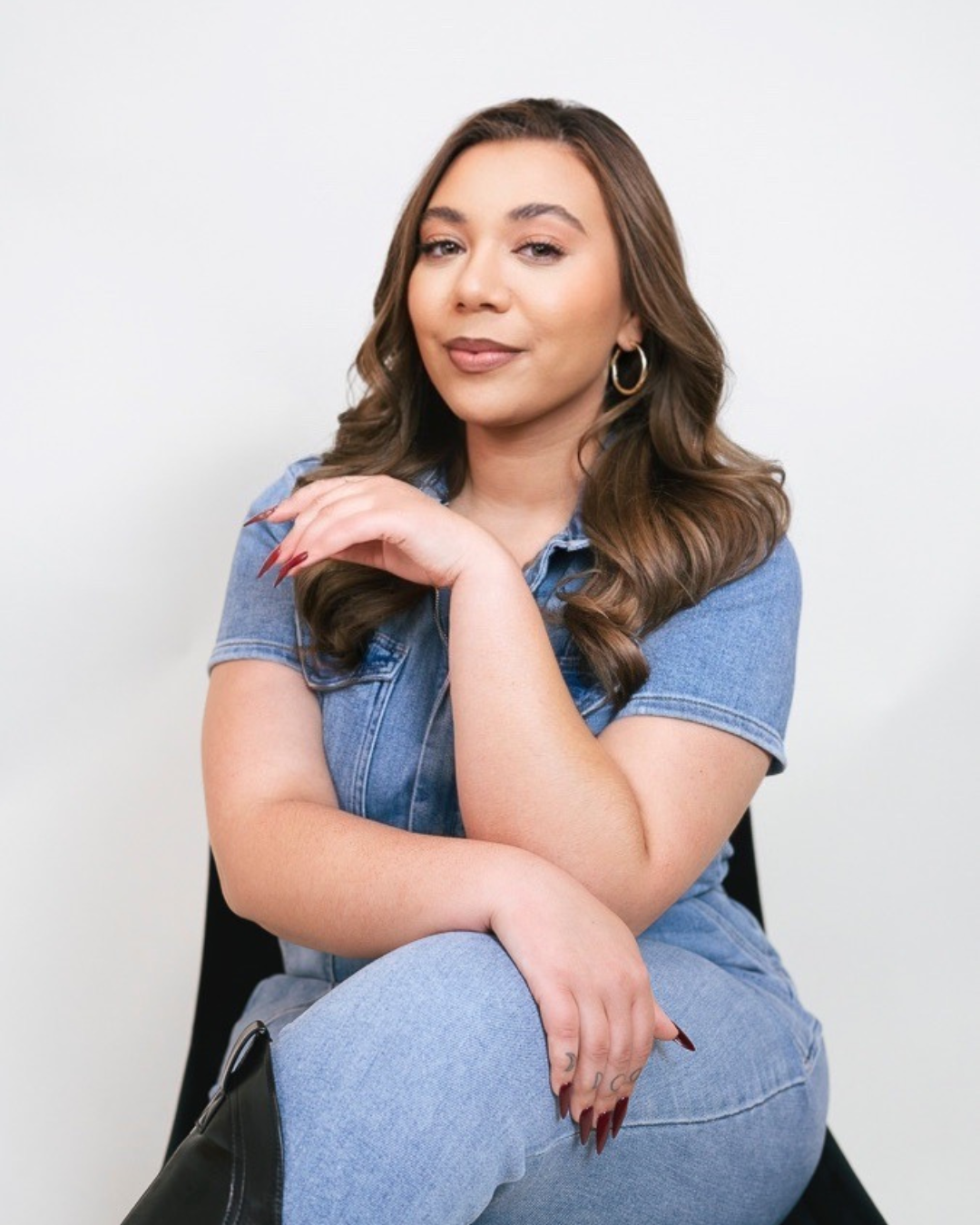 A woman with long brown hair sits confidently, wearing a denim jumpsuit and hoop earrings, with one arm resting on her knee and a slight smile on her face, against a plain white background. Elle Hope