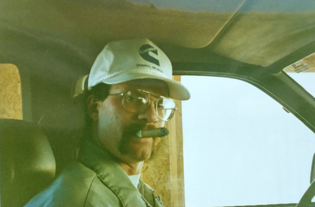 A man wearing a white cap and glasses sits in the drivers seat of a vehicle, looking at the camera with a cigar in his mouth. The background shows part of the cars interior and a wooden panel outside the window.