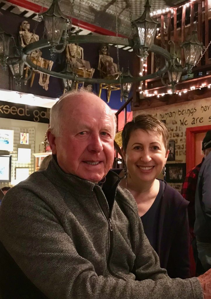 An older man and a younger woman smile at the camera inside a cozy, warmly lit bar decorated with hanging lanterns, skeleton figures, and string lights.