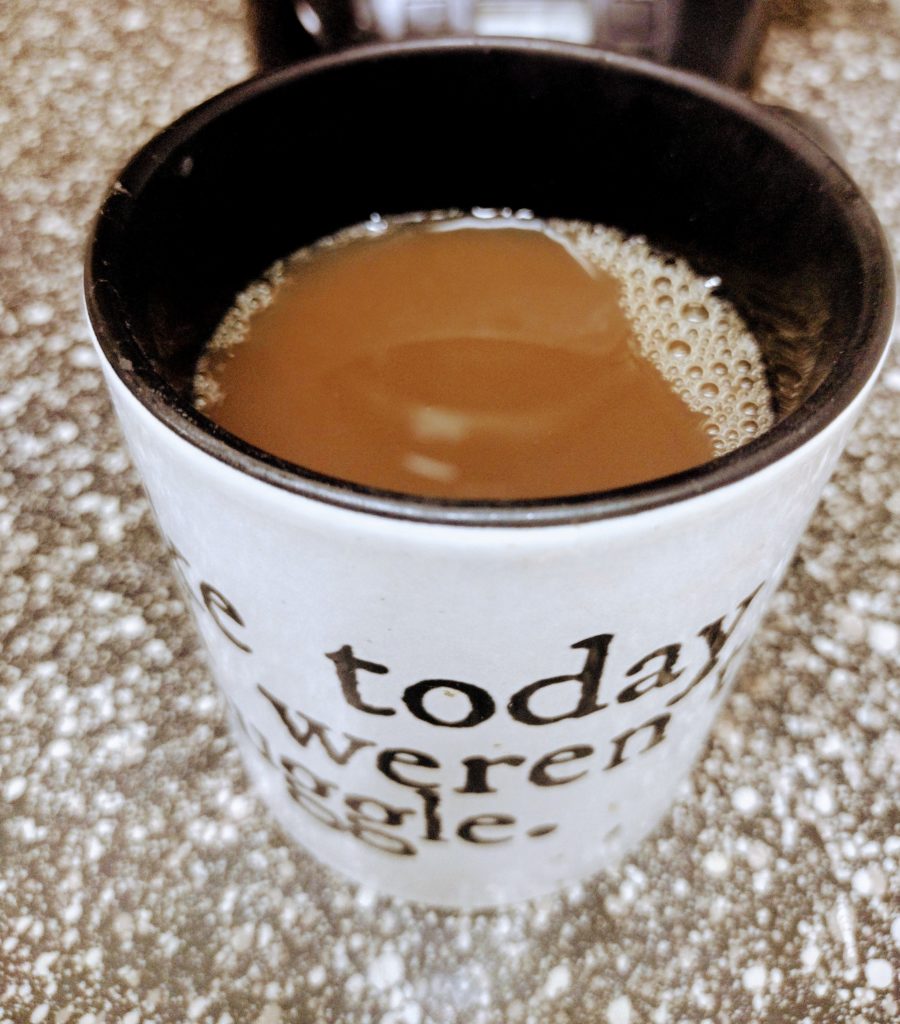 A white mug filled with coffee sits on a speckled countertop. The mug has black text on it, though only parts of the words are visible. Some bubbles are floating on the coffee’s surface.