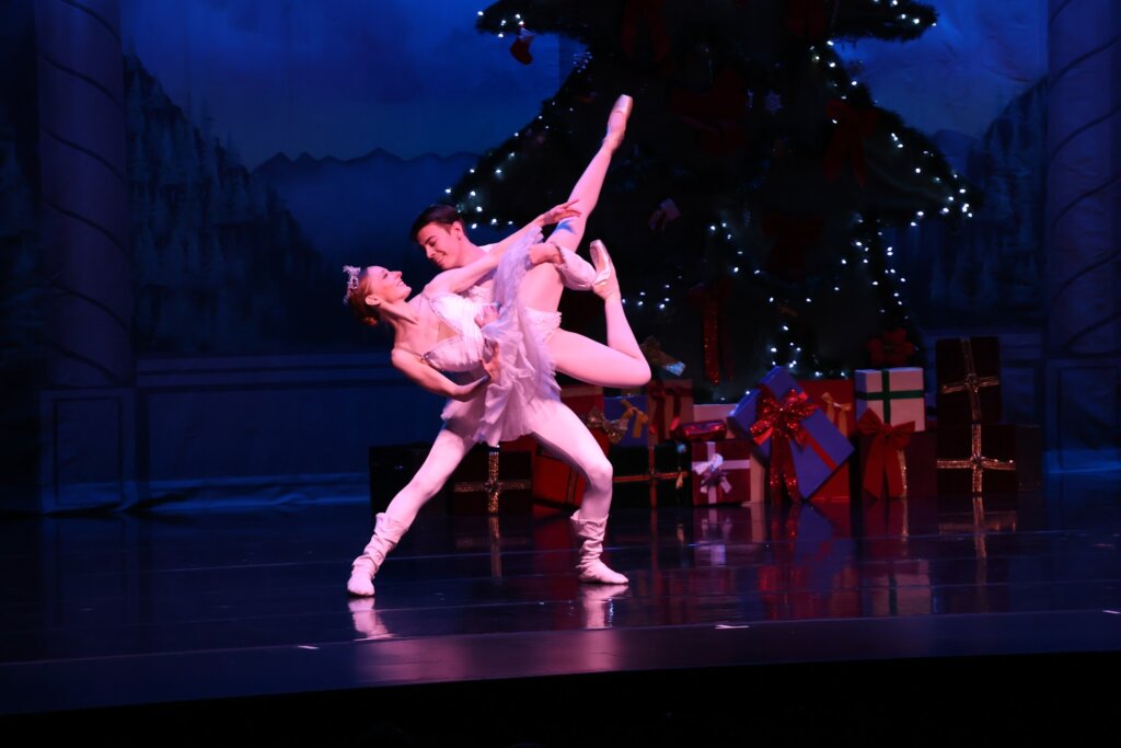 Two ballet dancers hold a pose in front of a holiday tree with presents.