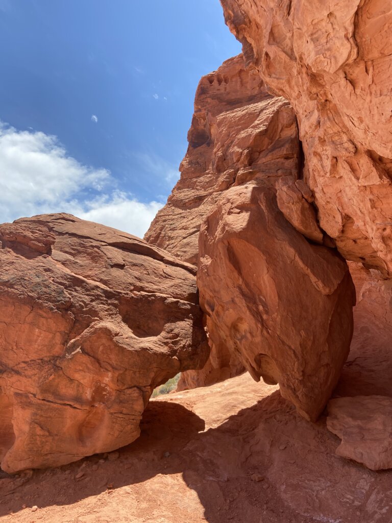 Large red sandstone rocks form a natural archway under a bright blue sky with some clouds. Sunlight casts shadows on the rugged, textured rock surfaces.