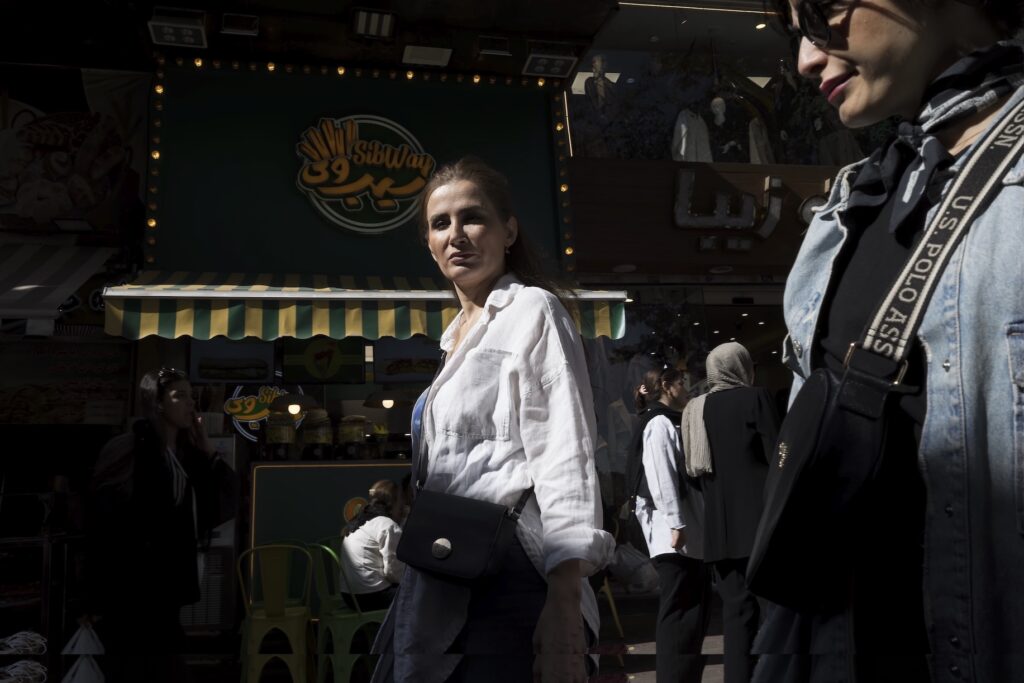 A woman in a white shirt stands in front of a storefront as other people walk by on a sunny street.