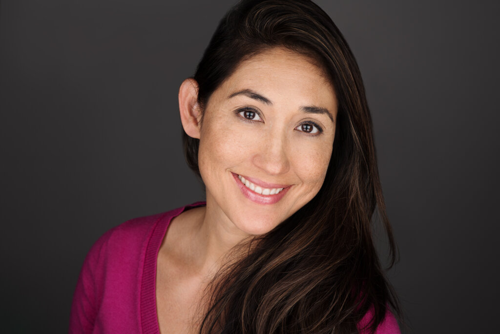 A woman with long dark hair smiles warmly at the camera. She is wearing a magenta sweater and stands against a plain dark gray background.