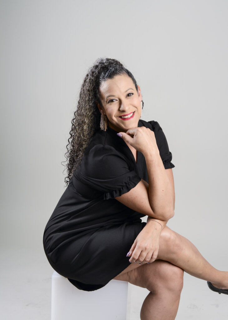 A smiling woman with curly hair, wearing a black dress and earrings, sits on a white cube against a light gray background, resting her chin on her hand and crossing her legs.