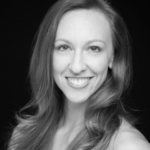 Black and white portrait of a young woman with long, wavy hair, smiling warmly at the camera against a dark background. She is facing forward, with her shoulders slightly angled.