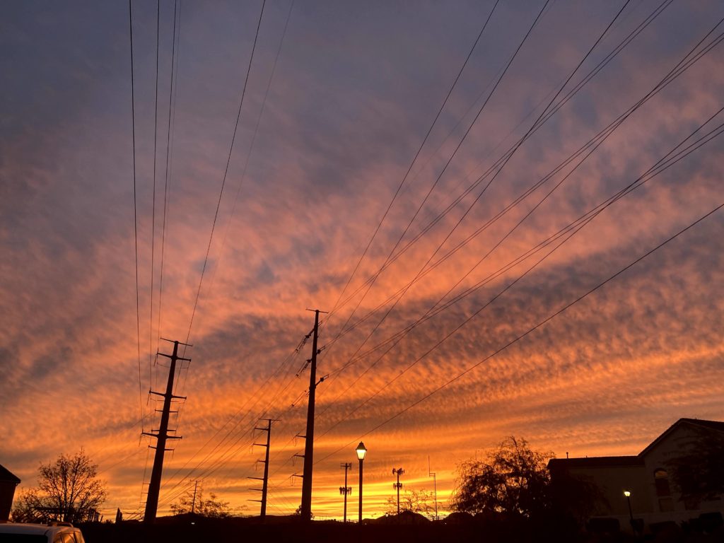 A vibrant sunset sky with orange, yellow, and purple hues, silhouetted power lines and poles, houses, trees, and streetlights in the foreground.