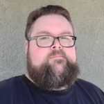 A person with short brown hair, a full beard, and glasses is standing in front of a textured beige wall, wearing a dark T-shirt and looking directly at the camera with a neutral expression.