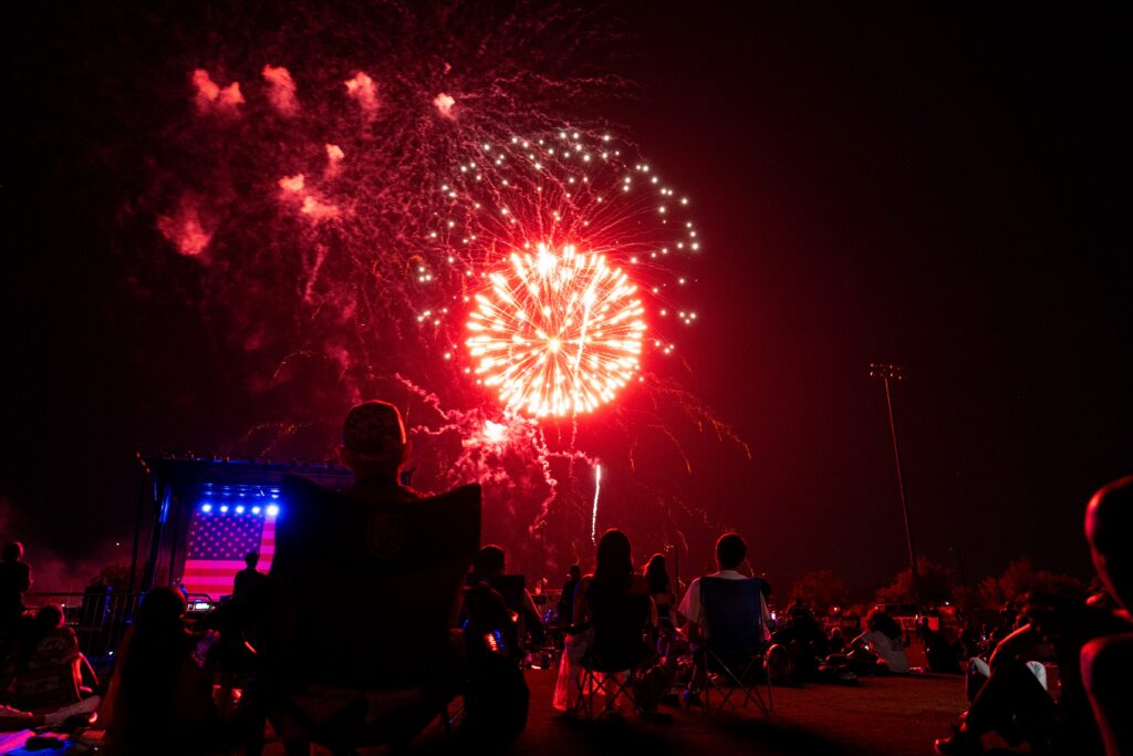 People sit outdoors at night watching bright red fireworks explode in the sky. Some are in lawn chairs, silhouetted against the glowing fireworks. A stage with blue and red lights is visible on the left side of the image.