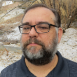 A middle-aged man with glasses and a salt-and-pepper beard stands outdoors in front of a snowy, leaf-strewn hillside. He is wearing a dark polo shirt and looks directly at the camera with a neutral expression.