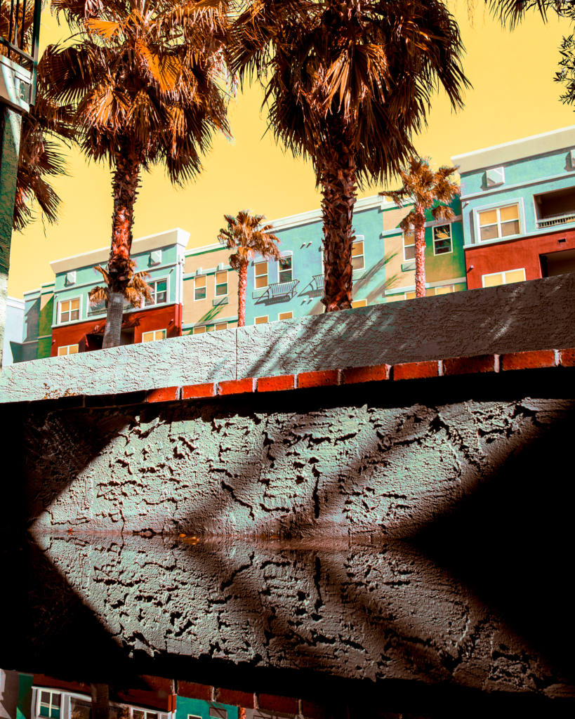 A colorful apartment building with palm trees in front, under a yellow sky. Shadows from a nearby structure create geometric patterns on a textured wall in the foreground.