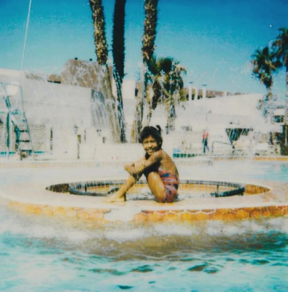 A smiling child in swim trunks sits on a circular platform in the middle of a pool, surrounded by splashing water. Palm trees and white buildings are visible in the sunny background.