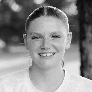 A young person with light skin and straight hair pulled back smiles at the camera. The background is blurred with hints of trees and sunlight. The image is black and white.