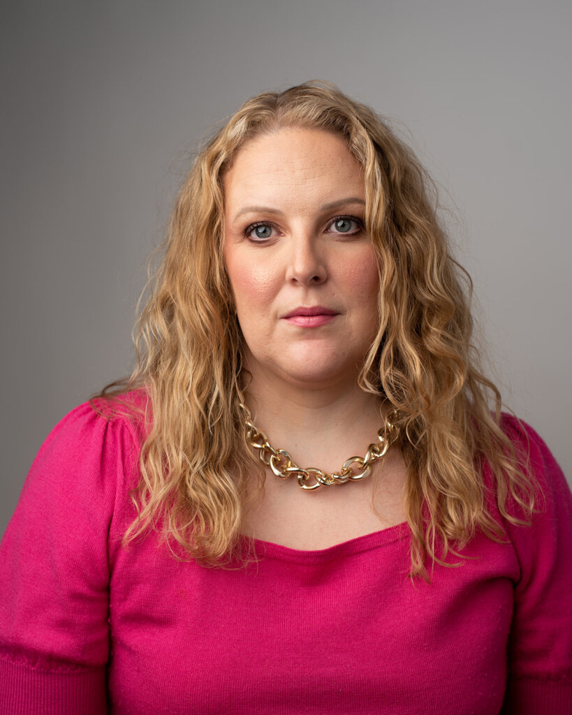 A woman with wavy blonde hair wearing a bright pink top and a chunky gold necklace poses against a plain gray background, looking directly at the camera with a neutral expression.