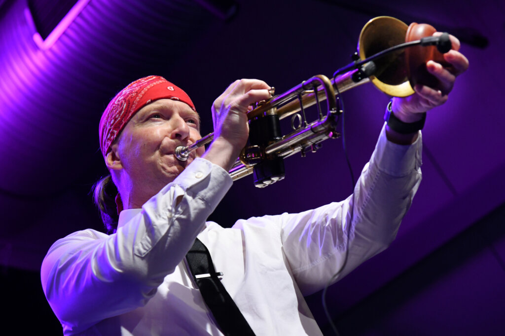A musician wearing a red bandana and white shirt plays a trumpet on stage under purple lighting.