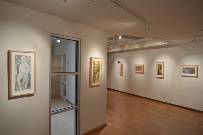 A brightly lit art gallery with wooden floors and cream walls displays several framed artworks.