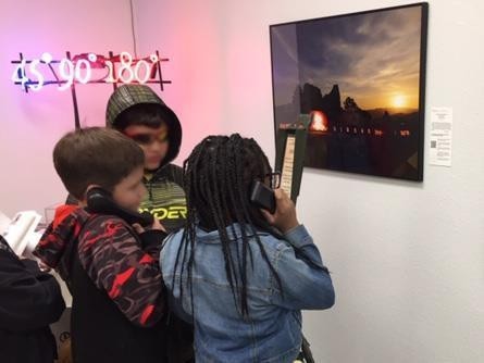 Three children listen to phones in an art gallery, viewing a framed sunset photograph.