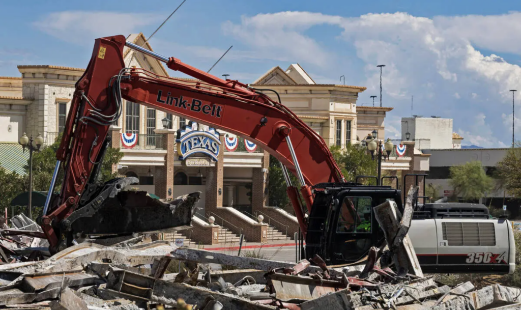 A red excavator demolishes a pile of concrete debris in front of a historic building decorated with American flags and a sign reading Texas. The sky is partly cloudy.