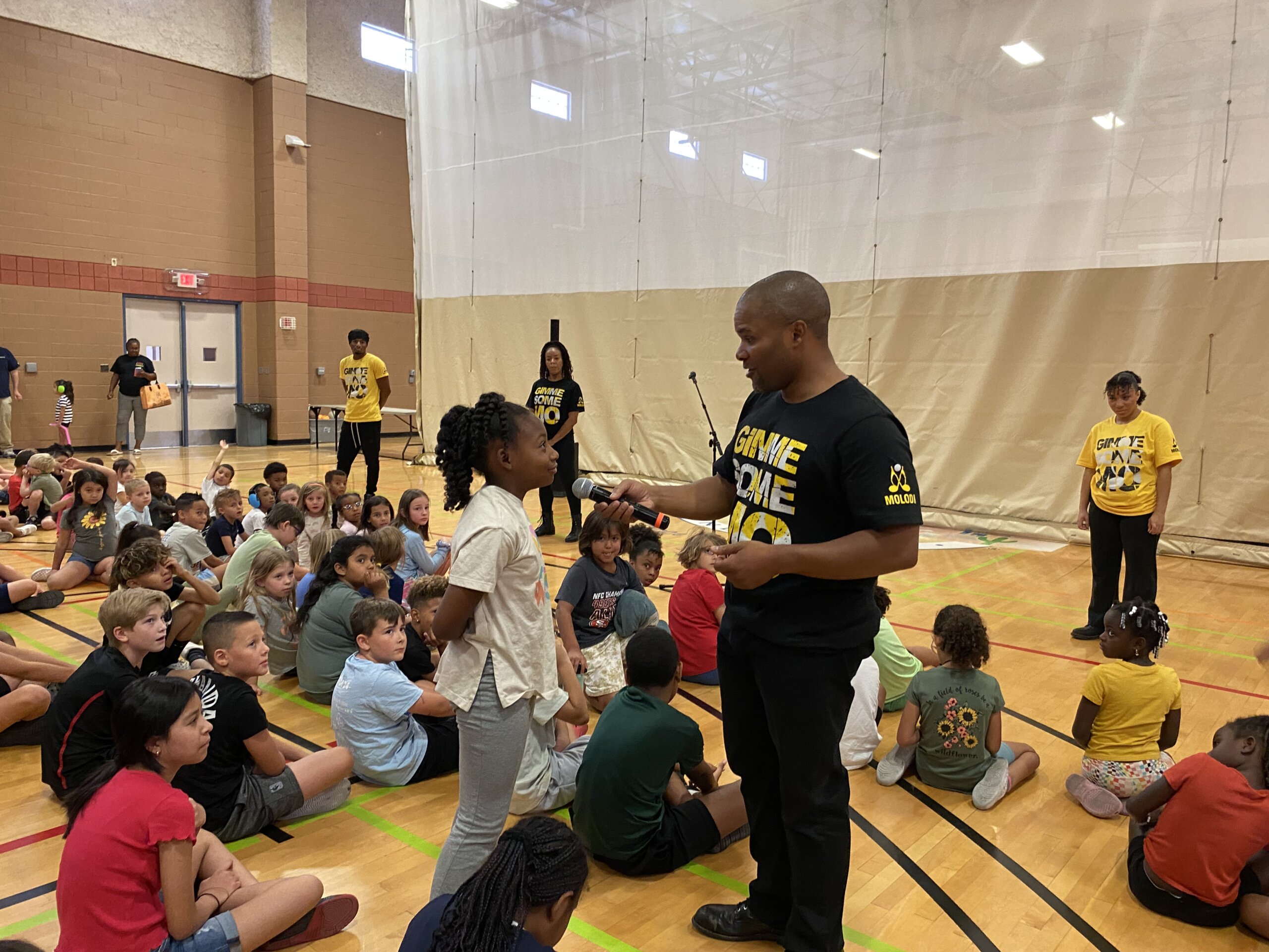 Jason Nious holds a microphone, talking to a young girl in a gymnasium. Children sit on the floor listening. Several adults stand in the background wearing matching "Gimme Some" shirts.