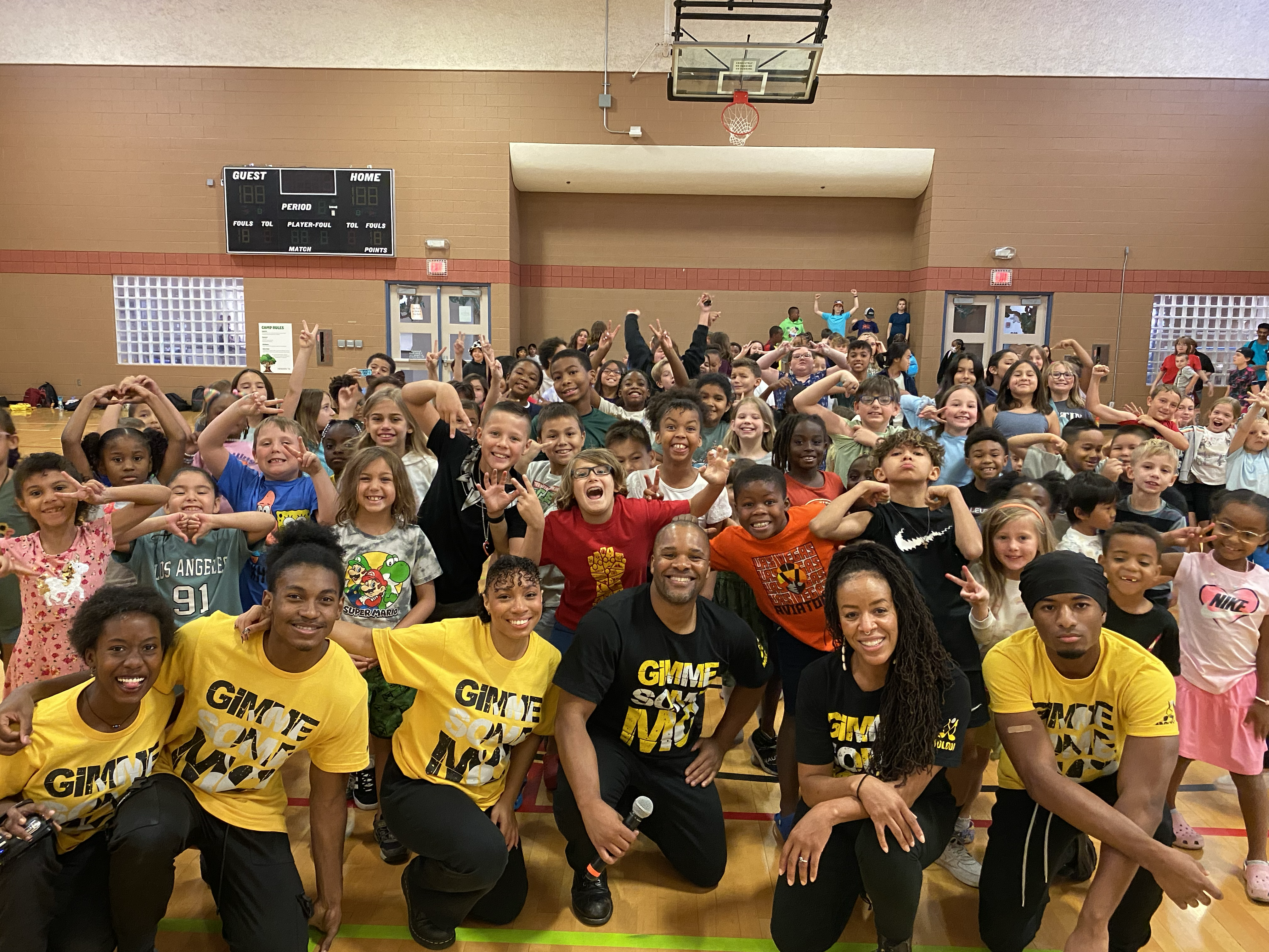 A diverse group of enthusiastic children and teachers are gathered in a school gym. Smiling widely, they strike playful poses, creating a joyful, lively atmosphere.