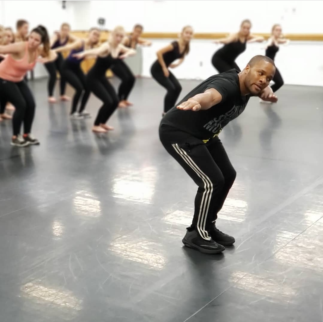 Jason Nious in a black outfit leads a group of students in a mirrored studio. They are all crouched, balancing with outstretched arms, conveying focus and energy.